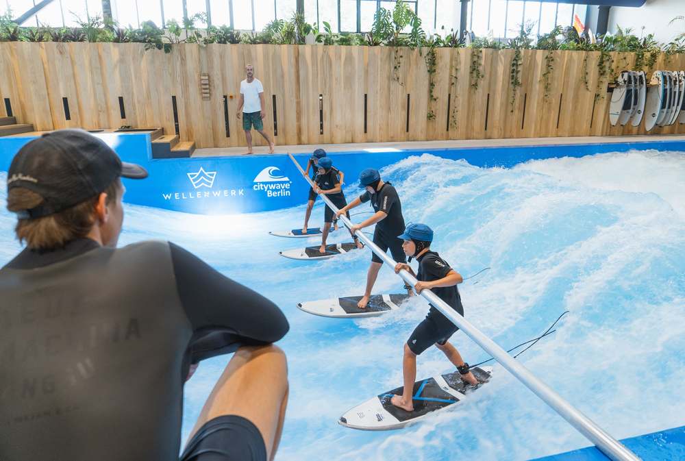 Kids camp group practices together with a support bar on the wave